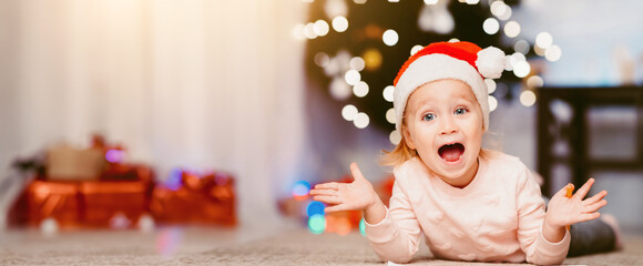 Christmas surprise. Happy emotional little girl in Santa hat lying on floor at home, opened mouth...