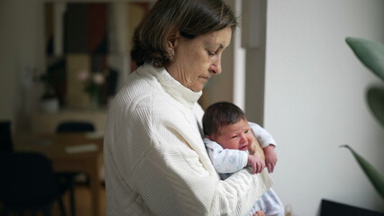 Obraz premium Elderly woman gently cradling a newborn baby, standing by a window in a softly lit room, embodying tenderness and care as she looks down at the infant with a calm and nurturing expression