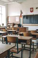 An empty vintage wooden classroom. There are lecture desks with chairs. The room has a chalkboard and windows. 