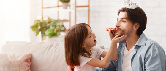 Small girl putting lipstick on daddy lips at home, dad wearing crown, copy space