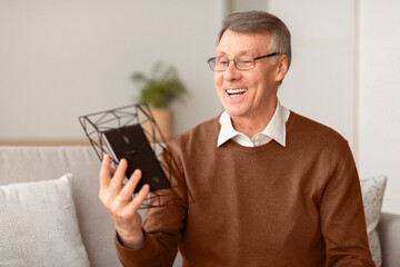 Happy Memories. Smiling Senior Man Looking At Family Portrait Holding Photo Frame Sitting On Sofa At Home. Selective Focus