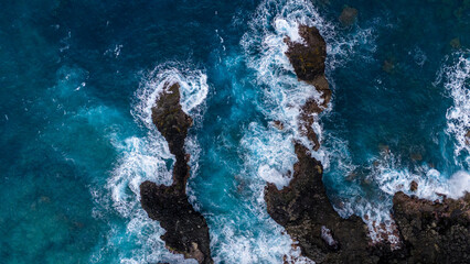 Overhead shot of sea waves flowing around isolated rocky outcrops, showcasing the contrasting hues of deep blue ocean water and jagged rocks.