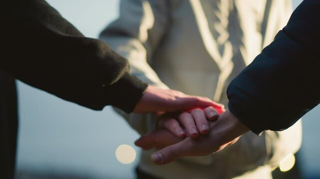 Close-up of three individuals stacking their hands together in a gesture of unity and teamwork, with a blurred background that includes someone wearing a gray jacket