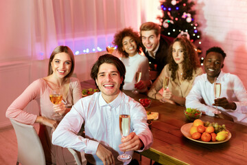 Happy New Year. Millennial Friends Holding Champagne Glasses Toasting Looking At Camera Sitting At Dinner Table At Home.
