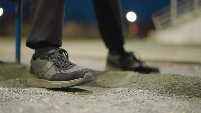 Close-up of a person's feet as they repeatedly tap on the ground, with a blurred, dimly lit background and an iron railing visible in the distance