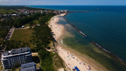 Aerial shot of Darłowo beach, showing golden sand, calm waters, beachgoers, and ships sailing from the port.