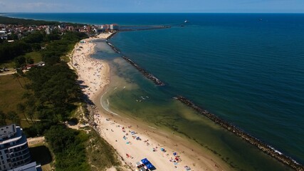 Drone captures Darłowo’s golden beach, sunbathers, swimmers, and ships heading out of the port into the calm Baltic Sea.