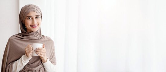 Relaxed arabic woman in headscarf standing with cup of coffee near window at home and looking at camera, enjoying weekend, empty space
