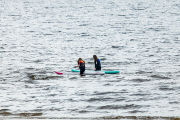 Obraz premium Two female paddle boarders on boards in sea water with island with lighthouse and hills on horizon