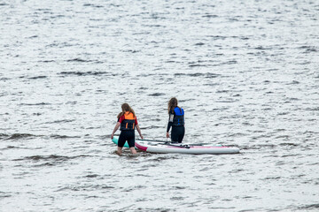 Obraz premium Two female paddle boarders on boards in sea water with island with lighthouse and hills on horizon