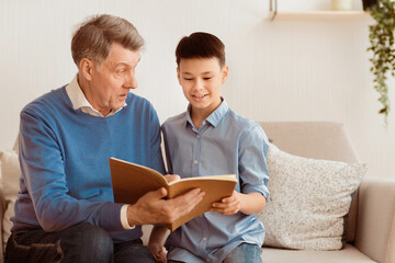 Fototapeta premium Elderly Grandfather And Grandson Boy Reading Book Sitting Together On Sofa At Home. Family Weekend. Selective Focus