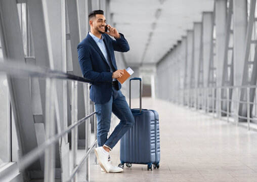 Mobile Call. Smiling Young Middle Eastern Businessman Talking On Cellphone In Airport While Waiting With Suitcase For Flight At Terminal, Happy Arab Man Having Pleasant Phone Conversation
