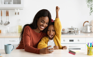 Happy Excited Black Mother And Little Daughter Celebrating Success With Smartphone While Sitting At...