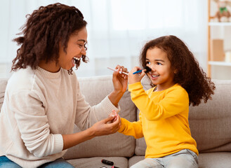 Black Mother And Little Daughter Making Makeup Together Having Fun Sitting On Sofa Indoor. Family Moments