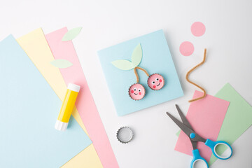 Flat lay of craft materials on white surface: pink, blue, green paper, blue-handled scissors, yellow glue stick, pink cherry cut outs with faces, green leaf on light blue square.