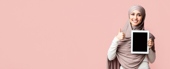 Cool app. Beautiful muslim girl in hijab holding digital tablet with black screen and gesturing thumb up over pink background with free space