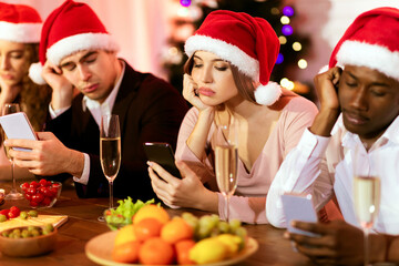 Bored Millennial Friends Using Mobile Phones During Dull Christmas Party Sitting At Celebratory Table Indoor