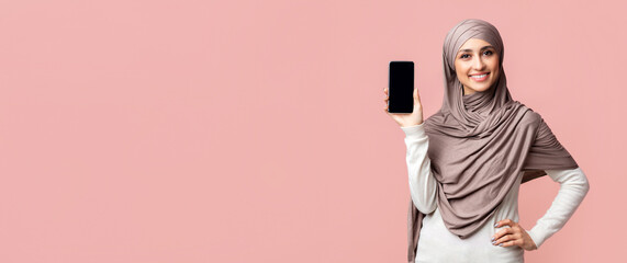 Happy arabic woman in hijab showing modern smartphone with black screen, standing over pink background in studio, free space