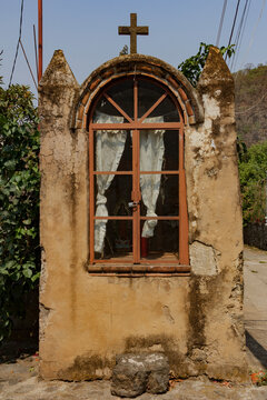 Antiguo nicho religioso r&uacute;stico con cruz, ventana arqueada y cortinas en una calle de un pueblo mexicano