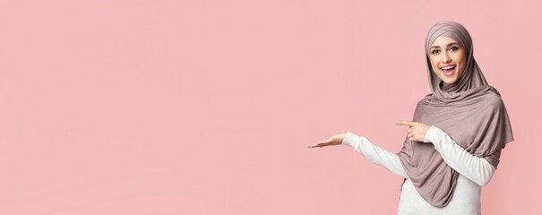 Beautiful muslim woman in hijab demonstrate something on her empty palm and pointing at it, standing over pink background, panorama with copy space