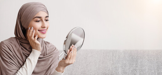 Beauty routine. Attractive muslim girl in hijab holding mirror and looking at her face with perfect...