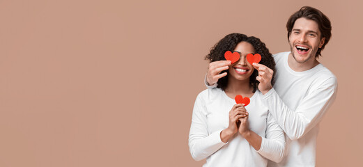 Falling In Love. Cheerful Man Holding Red Paper Hearts Over Eyes Of His Afro Girlfriend And Smiling At Camera, Yellow Background With Empty Space