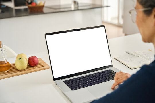 Middle aged woman customer looking at white mock up blank screen using laptop computer at home table doing online shopping, searching on web. Website mockup display template. Over shoulder view.