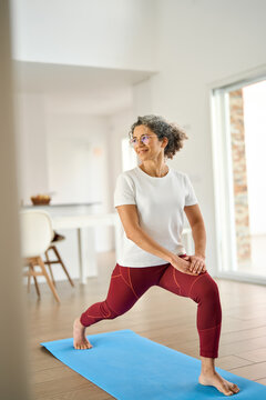 Sporty fit middle aged woman standing on mat at home doing fitness exercises morning routine. Happy healthy mature 50 years old lady enjoying casual workout or yoga in living room, vertical photo.
