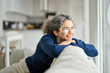 Happy middle aged woman sitting on comfy sofa in living room at home looking through window....