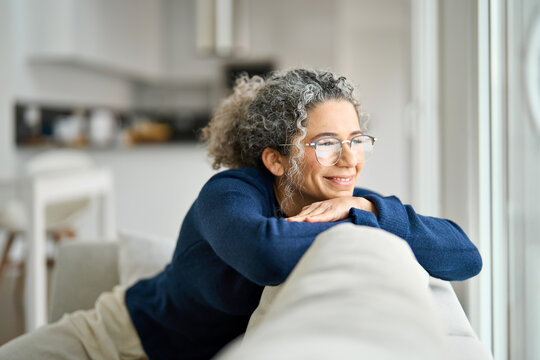 Happy middle aged woman sitting on comfy sofa in living room at home looking through window. Smiling mature older lady wearing glasses relaxing on couch looking away in modern cozy house.