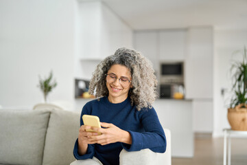 Happy curly middle aged older woman using phone sitting on sofa at home. Smiling relaxed mature...