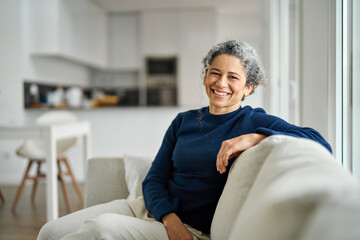 Happy middle aged woman sitting on comfy sofa in living room at home laughing. Smiling mature older...