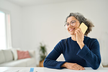 Happy middle aged woman making phone call sitting at table in living room. Smiling mature older...