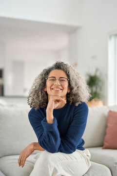 Vertical portrait of happy 50 years old woman sitting on sofa at home. Smiling middle aged lady looking at camera in living room at home. Mature older female with curly gray hair relaxing on couch.