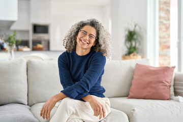 Happy middle aged woman in her 50s sitting on sofa in living room at home. Smiling beautiful mature...