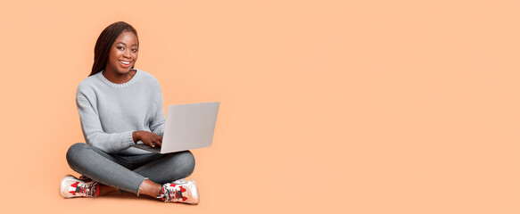 Dating website. Smiling young african woman sitting on floor and chatting online, using laptop, free space