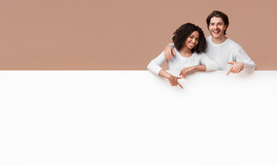 Place for your ad. Joyful interracial couple leaning and pointing at white advertisement board with...