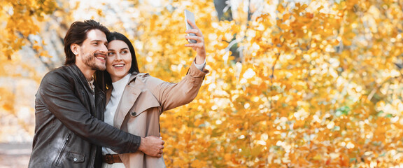 Cheerful young embraced couple in love taking selfie using smartphone at autumn park, copy space