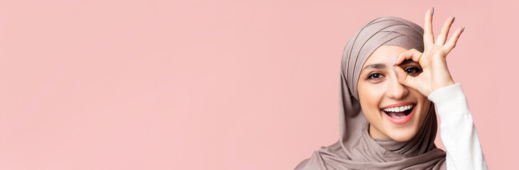 Cheerful arabic lady in hijab showing ok gesture, holding hand near eye, looking through fingers at camera, pink background with empty space, closeup