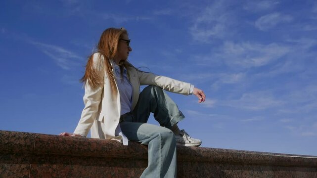 Thoughtful fashionable woman relaxing on parapet against blue sky. Young female with straight long hair is sitting on wall. She is spending leisure time during sunny day. 