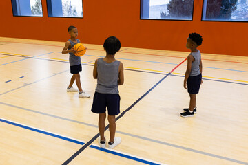 Fototapeta premium Playing basketball in school gym, three multiracial boys practicing together on court