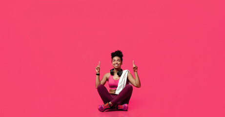 New ladies gym opening. Cheerful african american sporty girl with towel and earphones sitting on floor and pointing up over red background