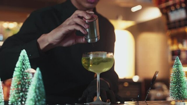 Closeup bartender putting cinnamon power on caramelized citrus slice on cube ice green Midori Sour cocktail served in stylish saucer glass at luxurious bar settings festive christmas theme. Vinosity.