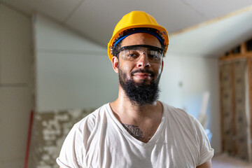 A cheerful male construction worker, in a bright safety helmet and glasses, smiles proudly at a busy remodeling site, showcasing his dedication to his craft among tools and materials