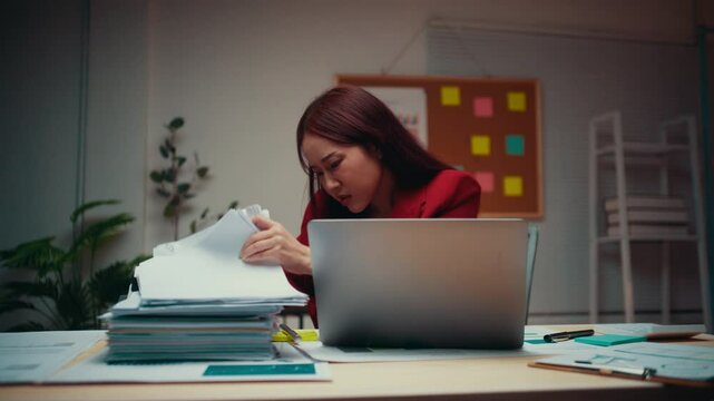 Asian accountant searches through a stack of paperwork on her desk, working late at night in her office. The atmosphere reflects the challenges of meeting deadlines and managing stress