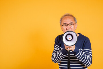 Portrait Asian old man wearing glasses screaming in megaphone studio shot isolated yellow...