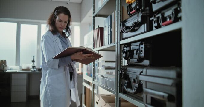 Female scientist or lab worker stands near shelf with folders, protective cases and boxes with fossils, reads scientific book and searches information for research. Modern archaeological laboratory.