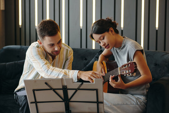 Professional instructor guiding student in playing guitar during music lesson in cozy room. Music stand and musical notes visible adding educational context to scene
