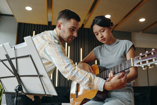 Guitar instructor demonstrating chords to engaged student in modern classroom setting. Both shown focusing attentively on guitar and music stand