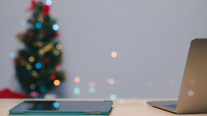 A cozy workspace featuring a laptop and tablet, with a blurred Christmas tree adorned in colorful...